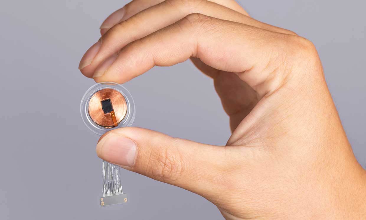  A close-up image of a Neuralink brain implant device, which is a small, round chip with a number of thin wires attached to it, being held by a person's thumb and forefinger.