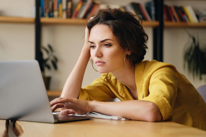 Young upset woman sitting at the desk tiredly working on new project with laptop in modern office